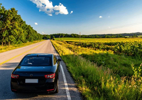 Black sedan parked along a rural roadside beside open fields under bright sunshine and blue skies.
