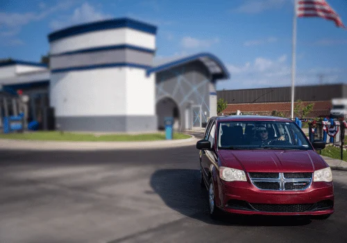 Red minivan arriving at a professional auto repair shop with a customer behind the wheel on a clear, sunny day.
